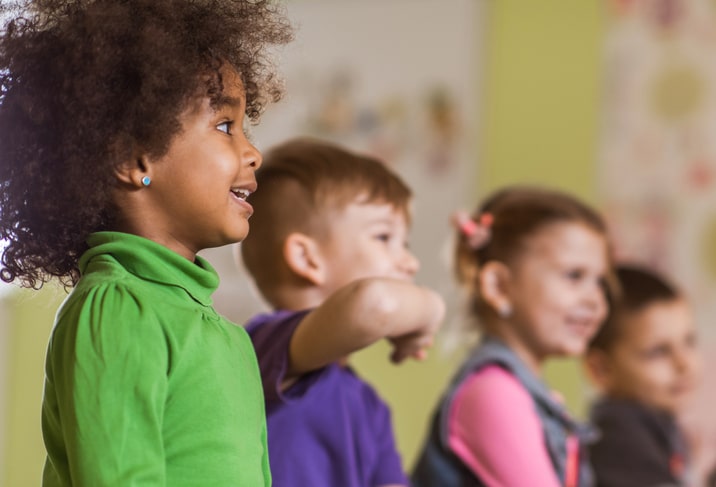 A black girl with a green shirt and curly hair stands next to three other children in a preschool