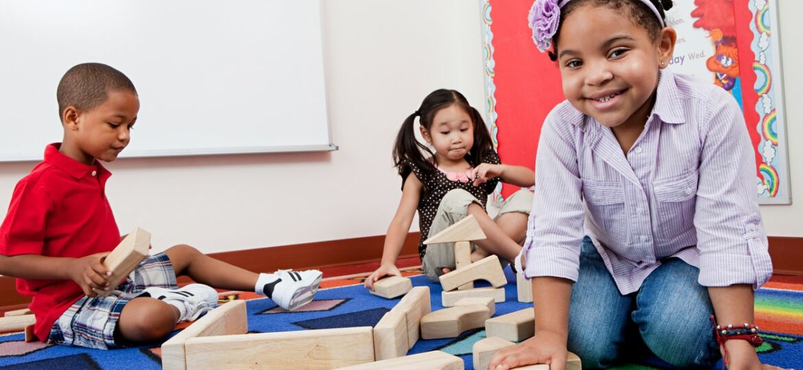 Three children sit on a rug playing together with building blocks