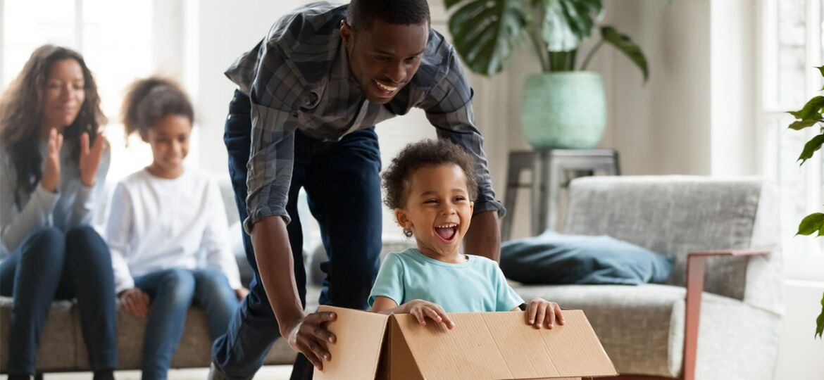 A boy in a blue shirt is pushed in a cardboard box by a man, with a young girl and woman sitting on a couch behind them