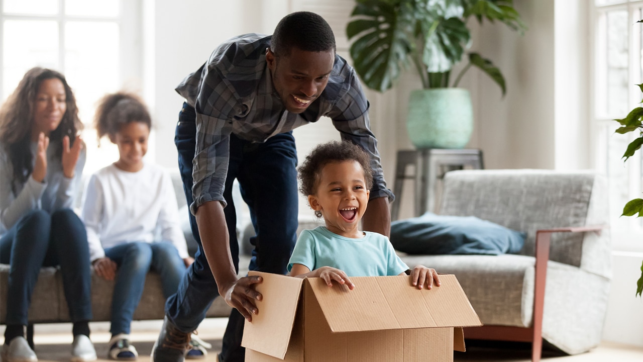 A boy in a blue shirt is pushed in a cardboard box by a man, with a young girl and woman sitting on a couch behind them