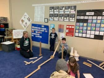 Two children build a block ramp while an adult watches and smiles.