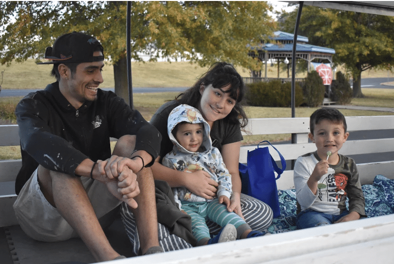 Two smiling adults and two small, smiling children sitting together on a wagon outside under trees.