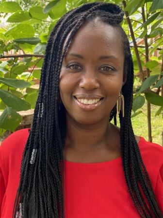 A smiling woman with braided hair, wearing a red top and earrings, standing in front of green foliage.