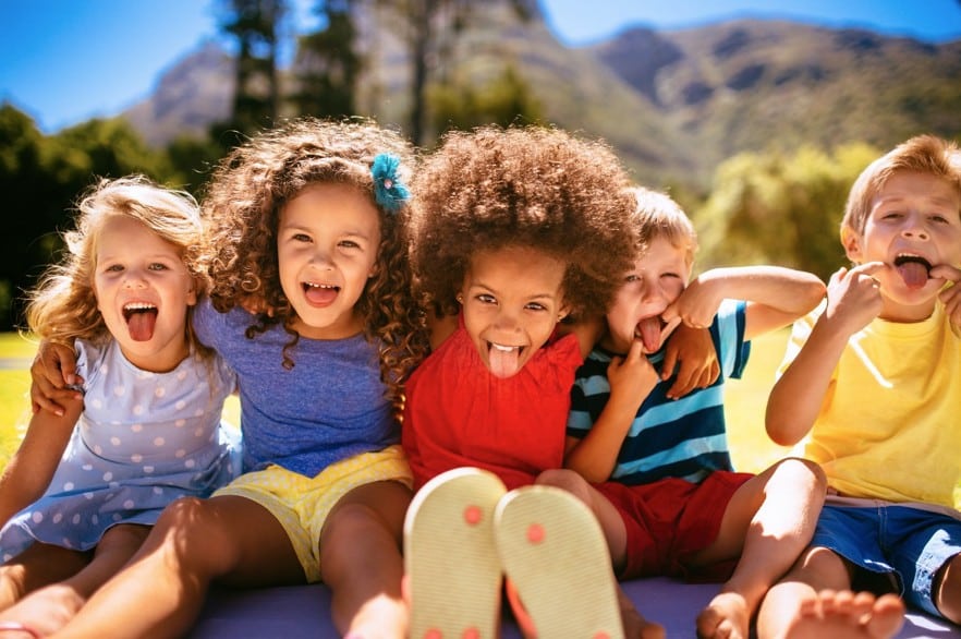 Five children sitting outdoors in a row, smiling and sticking out their tongues, with mountains and trees in the background on a sunny day.
