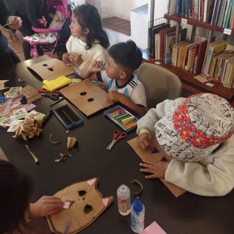 Three children sit at a table creating crafts with cardboard, markers, and glue; books are visible on shelves in the background.