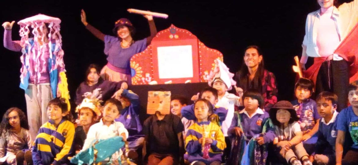 A group of children and adults onstage in costume, holding props and colorful decorations, pose together for a group photo against a black background.