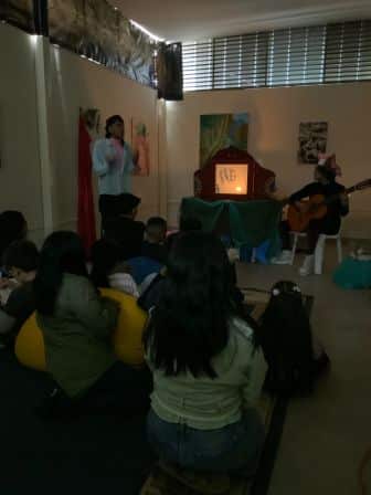 Several children sit on the floor facing a woman telling a story near a lit puppet theater, while another person plays guitar.