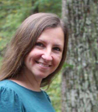 A woman with long brown hair and a teal top smiles outdoors in front of a tree and greenery.