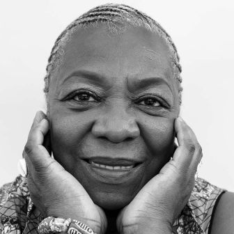 Elderly woman with braided hair smiles softly, resting her face in her hands. She wears rings and beaded bracelets. The photo is black and white with a plain background.