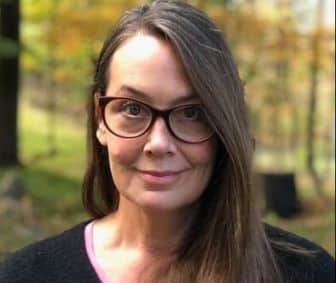 A woman with long brown hair and glasses stands outdoors, facing the camera, with blurred green and yellow foliage in the background.