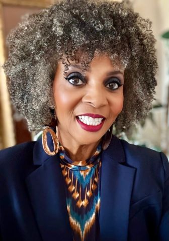 A woman with curly gray hair, wearing a navy blazer, patterned top, and large hoop earrings, smiling at the camera.
