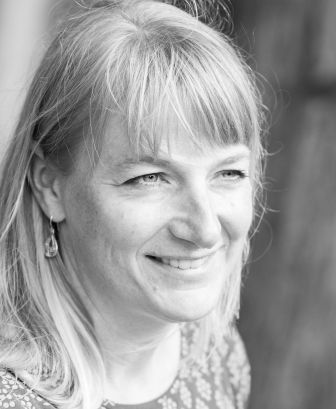 A woman with light-colored hair and earrings is smiling, looking slightly to the right. The image is in black and white.