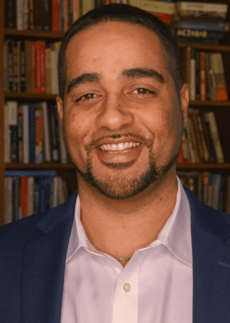 A man in a suit and collared shirt smiles in front of a bookshelf filled with books.