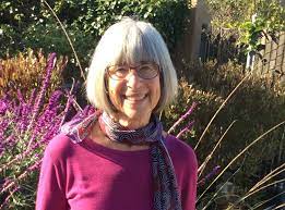 An older woman with gray hair and glasses, wearing a purple top and patterned scarf, stands outside in front of purple flowering plants and greenery.