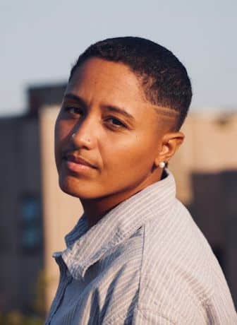 Person with short hair and two nose piercings wearing a striped button-up shirt, standing outdoors with a building in the background.