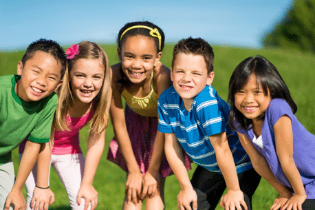 Five children stand outdoors on grass, smiling at the camera on a sunny day. They are close together and appear happy.