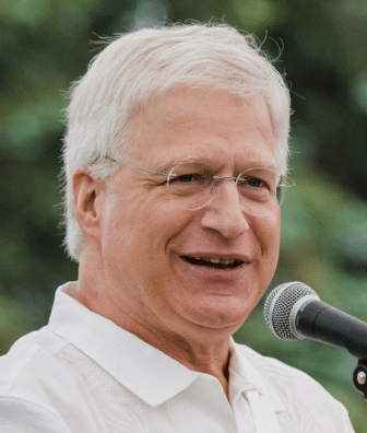 An older man with white hair and glasses speaks into a microphone outdoors, wearing a white collared shirt.
