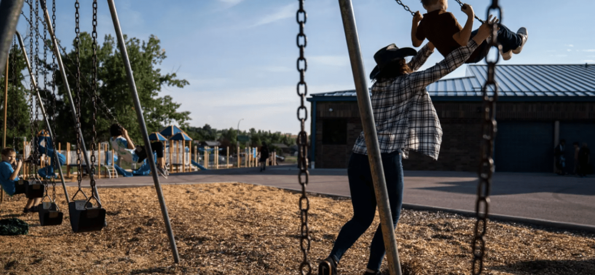 A woman pushes a young boy on a swing at a playground under a clear sky, with empty swings and a building in the background.