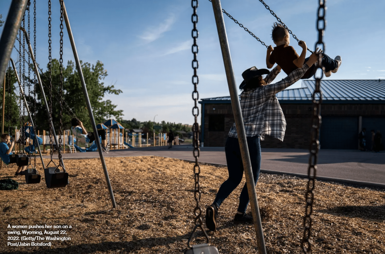 A woman pushes a young boy on a swing at a playground under a clear sky, with empty swings and a building in the background.