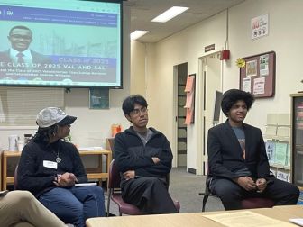 Three students sit in chairs during an indoor discussion, with one person wearing a suit. A projection screen and office materials are visible in the background.