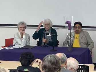 Three older women sit at a table in front of an audience; one speaks into a microphone, another listens, and a potted plant is on the table.