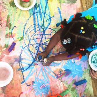 A child with colorful hair accessories draws on a large paper with blue chalk, surrounded by art supplies and vibrant scribbles.