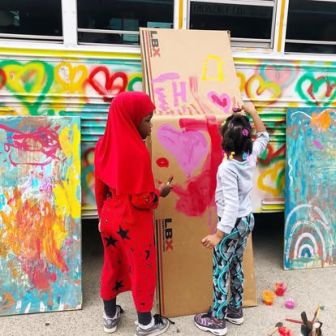 Two children paint colorful hearts on a cardboard panel in front of a bus decorated with heart designs and painted art boards.