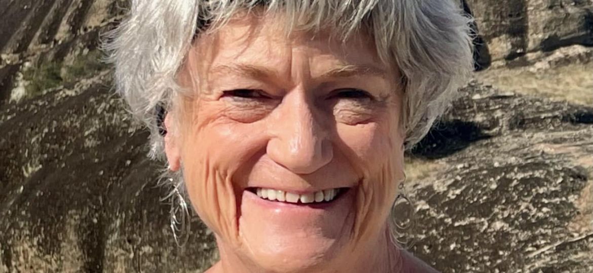 Older woman with short gray hair and a floral top smiles outdoors with rocky hills and a large rock formation in the background.