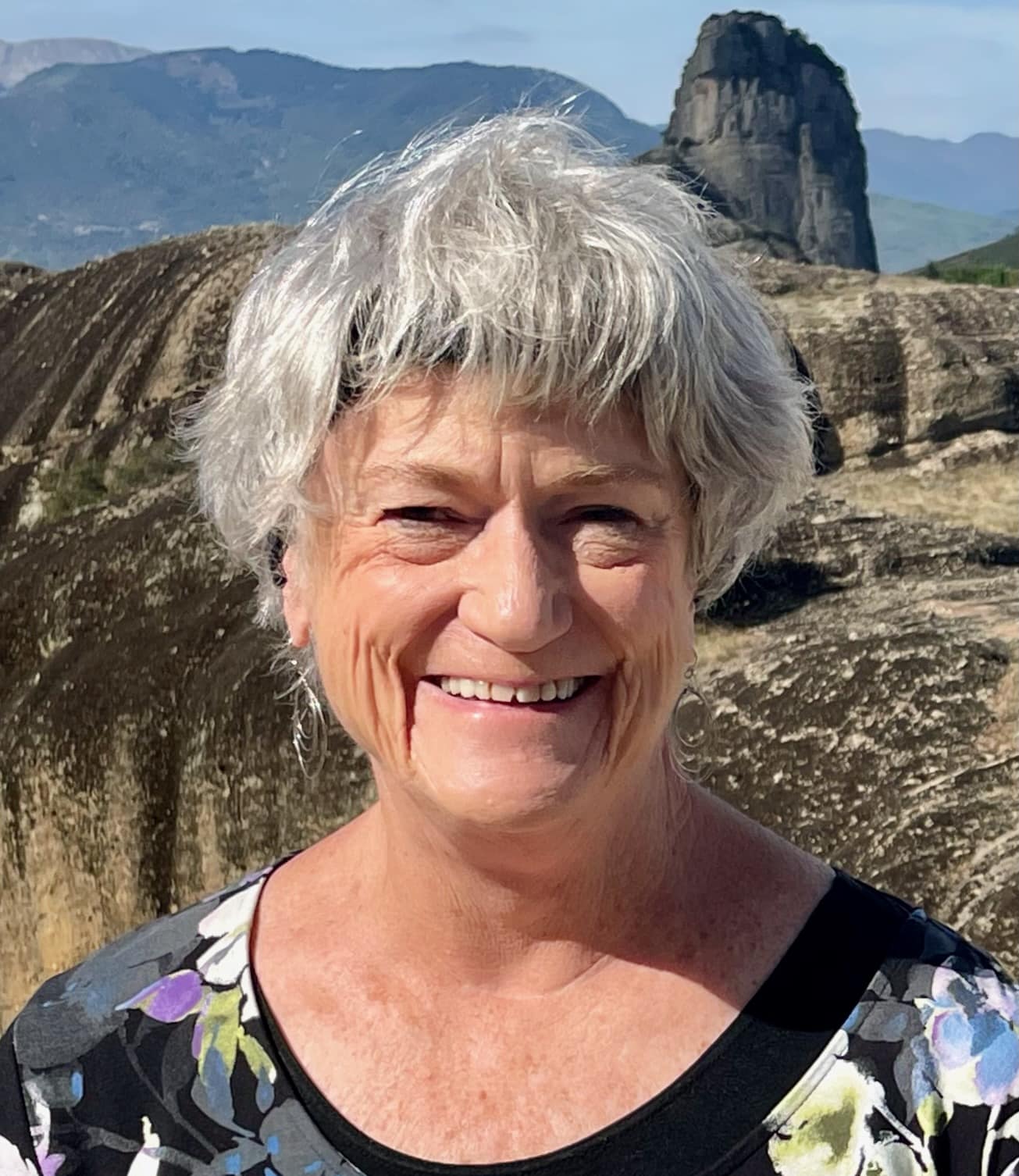 Older woman with short gray hair and a floral top smiles outdoors with rocky hills and a large rock formation in the background.