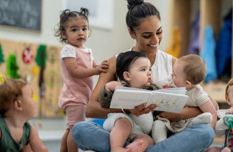 An adult sits on the floor reading a book to three babies in her lap while another toddler stands nearby in a daycare setting.