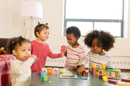 Four young children play with colorful blocks and toys on a table in a bright room with a window and a lamp in the background.