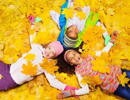Three children lying on autumn leaves, dressed in colorful winter clothes, smiling and holding yellow leaves.
