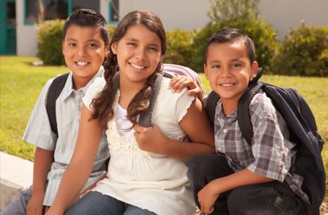 Three children with backpacks sit outdoors, smiling at the camera.