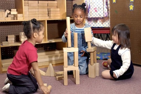 Three young children sit on the floor building a tower with wooden blocks in a classroom setting.