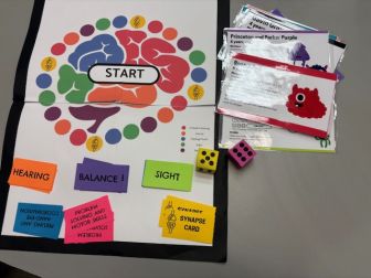 A colorful board game setup with a brain-themed board, cards labeled “HEARING,” “BALANCE,” “SIGHT,” two dice, and several instruction cards on a gray table.