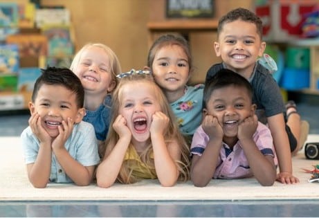 Six young children lying on a rug indoors, smiling and posing happily for the camera.
