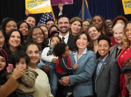 A diverse group of people, including adults and children, gather for a photo. Some hold "Child Care For All" signs, and one woman in front is holding a child.
