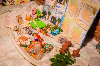 A display of handmade clay animal figures and drawings labeled "Sunny" and "Windy" on a plastic sheet, with some leaves and small objects arranged around them.