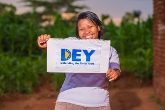 A person stands outdoors holding a sign that reads "DEY Defending the Early Years," with green foliage visible in the background.