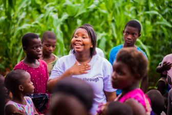 A woman smiling with her hand on her chest stands among a group of children outdoors in front of tall green plants.