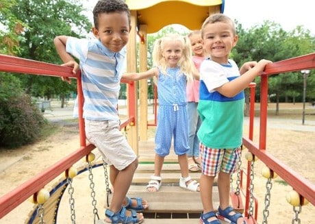 Four young children stand and smile on a playground bridge, surrounded by trees and playground equipment on a sunny day.