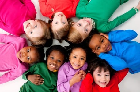 Nine children wearing colorful shirts lie on their backs in a circle on a white surface, smiling and looking up at the camera.