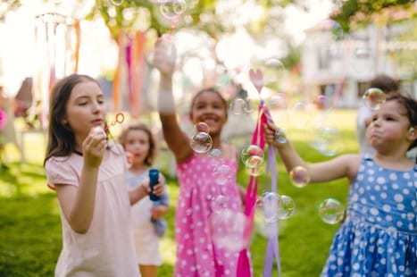 Several young children in colorful dresses blow and chase bubbles outside on a sunny day, with a blurred green background and decorations.