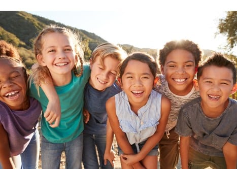 A group of six children stand outdoors in a row, smiling and laughing with their arms around each other, with trees and hills in the background.